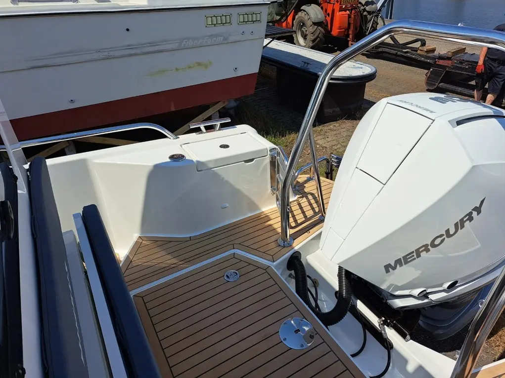A close-up of a boat's deck featuring a Mercury outboard motor, polished metal railing, and wooden flooring, with a second boat in the background.
